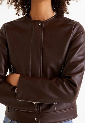 Person iført en mørkebrun læderjakke med lynlåslommer og ærmer, krydsede arme, der viser jakken forfra og teksturdetaljer.