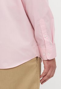 Camisa de manga larga rosa claro con una textura suave, puños abotonados y un dobladillo curvado, combinada con pantalones de color beige.