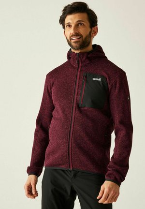 Mann trägt eine kastanienrote Fleecejacke mit Reißverschluss, schwarzer Brusttasche und Kapuze, steht vor einfachem hellem Hintergrund.