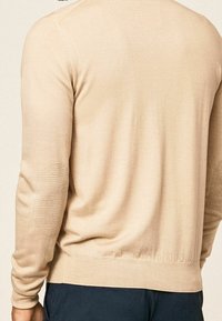 Pull beige à manches longues, ourlet et poignets côtelés, avec des coudières texturées en motif tricoté contrastant. Finition en tissu lisse.