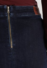 Dettaglio ravvicinato di una gonna di jeans blu scuro con una cerniera verticale in ottone e una patch in pelle marrone del brand vicino all'orlo della vita, indossata con un top bordeaux.