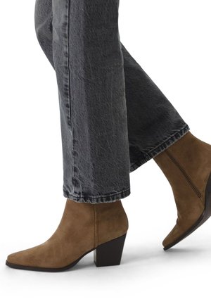 Bruine suede enkelboots met een puntige neus en een blokhak, gecombineerd met vervaagde zwarte wijde jeans. Ritsdetail zichtbaar aan de zijkant.