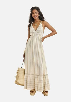 Mujer vistiendo un vestido maxi sin mangas crema con rayas, sandalias con plataforma, collar dorado, pulsera y llevando una bolsa de mano tejida con flecos.