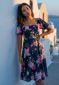 Frau mit dunklem Haar, die ein marineblaues Blumenkleid trägt und sich bei Sonnenuntergang an eine weiße Wand in der Nähe weißer Gebäude und eines Gewässers lehnt.