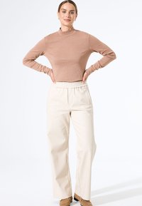 Langsleeve, fitted bronzen top met een hoge hals; gecombineerd met relaxte crèmekleurige broek met elastische taille. Neutralkleurige schoenen maken de outfit compleet.