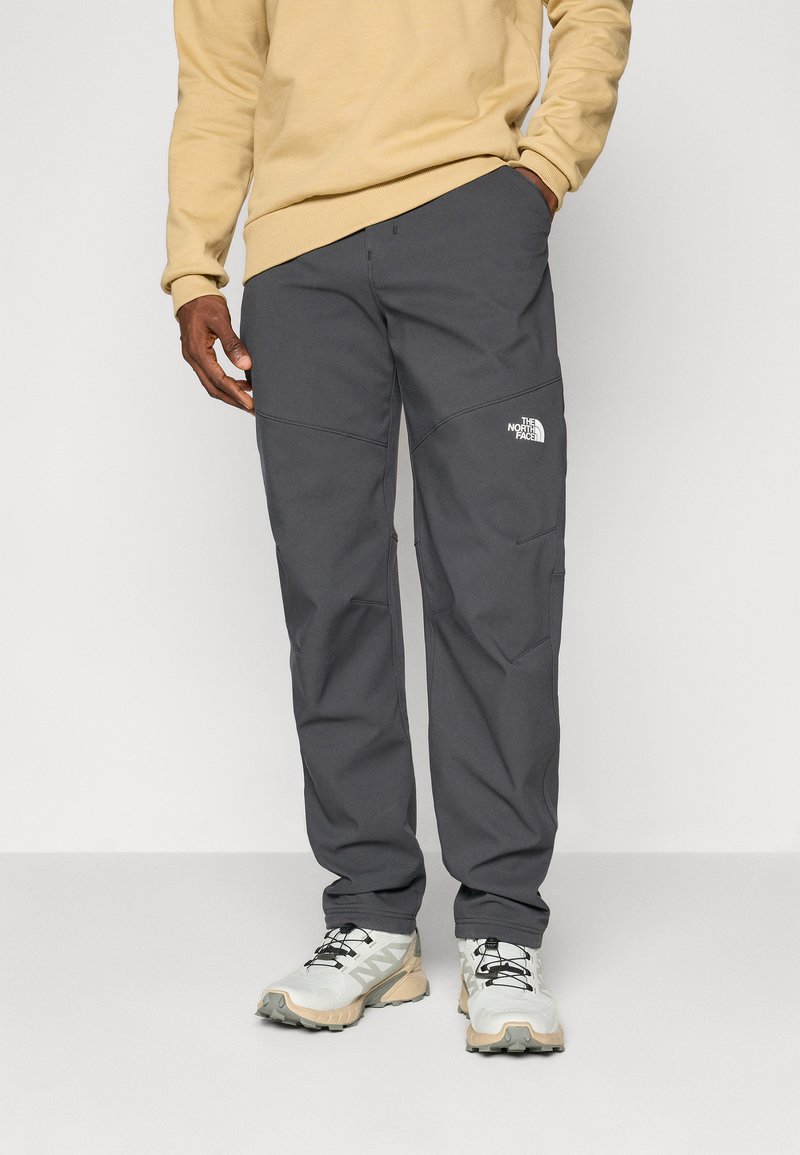 Grijze, lichtgewicht buitenbroek met kniestukken en het logo van The North Face. Draagt een beige sweater en lichte hardloopschoenen.
