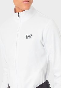 Chaqueta blanca con cremallera, fabricada en un tejido suave, que presenta un logo negro de EA7 en el lado izquierdo del pecho y un cuello alto. Diseño elegante y deportivo.