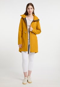 Giacca gialla impermeabile con colletto alto, due tasche frontali e chiusura con zip. Indossata sopra una maglietta a righe e leggings bianchi.