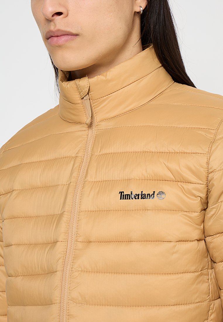 Giacca puffer beige con colletto alto, cuciture orizzontali e chiusura a zip. Presenta il logo Timberland sul lato sinistro del petto.