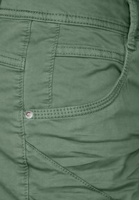Groene stof met een gladde textuur heeft een zijzak en contrasterende stiksels. Opvallende details zijn een metalen drukknoop en een diagonale naad.