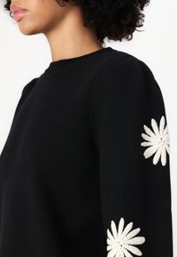 Svart genser med lange ermer, prydet med hvite broderte blomsterdesign på mansjettene. Rund halsdesign, myk tekstur.