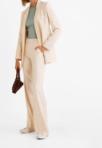 Beige tailored suit with wide-leg trousers and a fitted ribbed green top, complemented by a textured brown handbag and white footwear.
