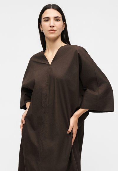 Γυναίκα με ίσια σκούρα μαλλιά φοράει αέρινο σκούρο καφέ φόρεμα με φαρδιά μανίκια, ποζάροντας με τα χέρια στις τσέπες μπροστά σε απλό φόντο.