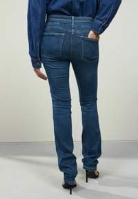 Sötétkék denim farmernadrág, slim fit szabással, öt zsebbel, megerősített varrásokkal és közepes derekú kivitelben, fekete stiletto cipővel párosítva.