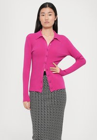 Taillierte Strickjacke aus leuchtend pinkem Material mit Kragen, langen Ärmeln und einer Knopfleiste, kombiniert mit einem gemusterten schwarzen Rock.