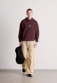 Felpa con cappuccio color marrone scuro con tasca frontale e toppa con logo, abbinata a pantaloni larghi color crema. La modella tiene una borsa rotonda nera, in piedi su una superficie chiara.