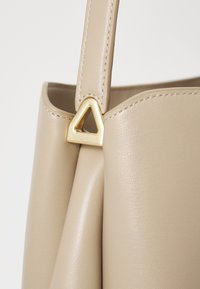 Beige leather bag with a structured design, featuring a triangular gold hardware accent on the strap and visible stitching details.