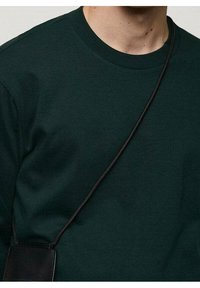 Personne portant un sweat-shirt vert foncé à col ras du cou avec une fine sangle noire traversant la poitrine, attachée à une pochette en cuir noir ou un petit sac.