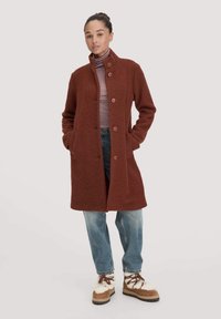 Cappotto di lana marrone con chiusura a bottone, due tasche laterali e colletto. Indossato sopra un dolcevita a righe e abbinato a jeans chiari e stivali.