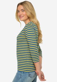 Donna bionda che indossa una maglietta a maniche lunghe blu e gialla a righe e jeans azzurri, sorridente e con lo sguardo rivolto di lato, con le mani dietro la schiena.