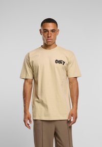 Mladý muž s krátkými vlasy a pihami, oblečený v béžovém tričku OBEY a světlých hnědých kalhotách, stojící před jednobarevným šedým pozadím.