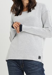 Top de punto de malla de manga larga en gris claro con puños en azul marino y un pequeño parche de logo en el dobladillo. Corte relajado, tejido texturizado, cuello redondo.