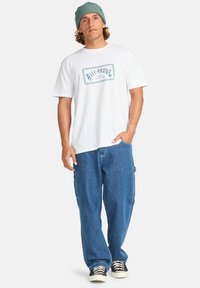 Hvid kortærmet t-shirt med blåt trykt logo, parret med løstsiddende blå denimbukser og sort-hvide stribede sneakers.