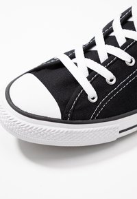 Zapatilla de lona negra con puntera de goma blanca, cordones blancos y costuras negras. La suela texturizada presenta un patrón de diamante para mayor agarre.