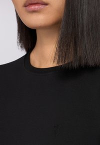 Černé bavlněné tričko s kulatým výstřihem a krátkými rukávy, s jemně vyraženým logem na hrudi. Hladká textura.