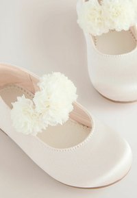 Valkoinen satiininen ballerinatossu, jossa on sileä pinta, varustettuna kahdella valkoisella kangaspompomilla remmissä ja pyöristetty kärki.