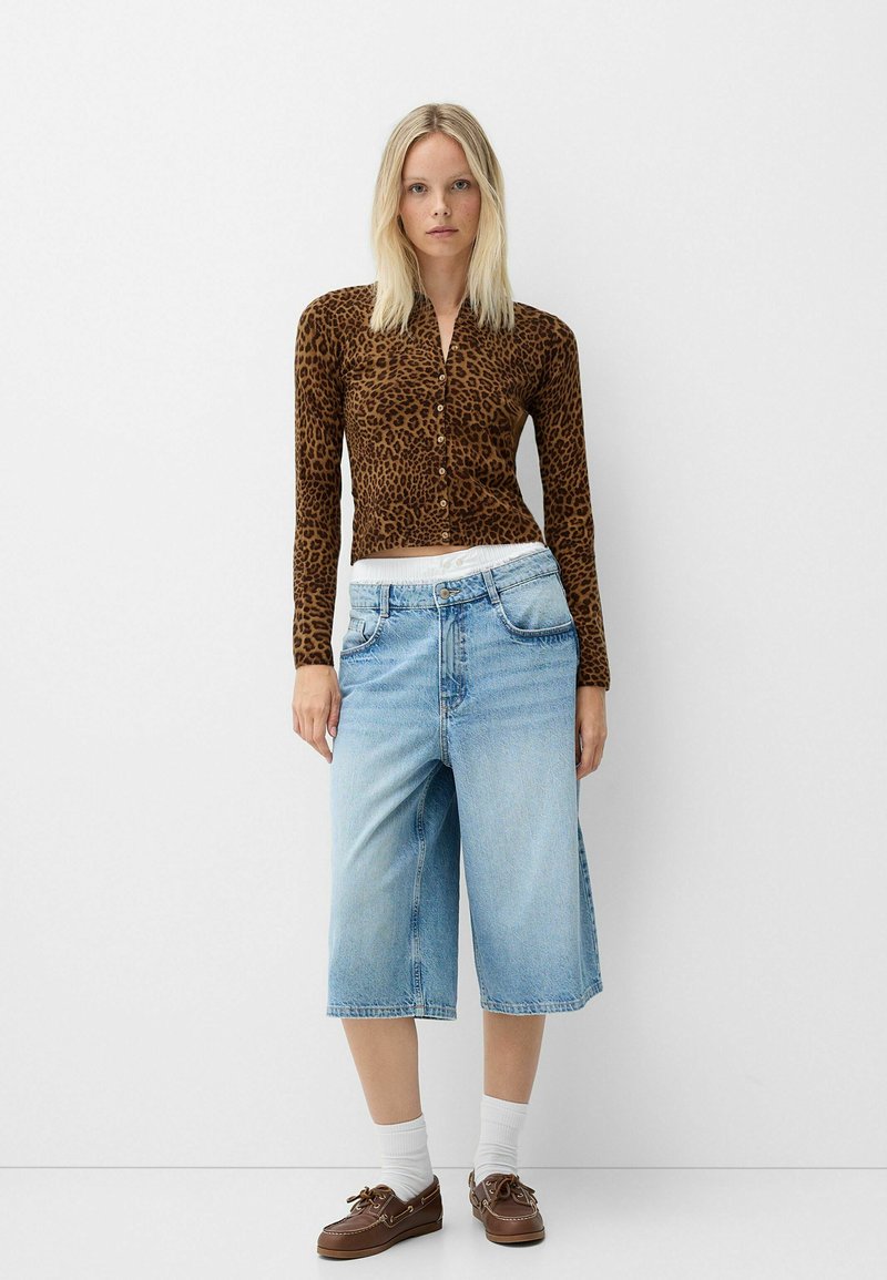 Braun geflecktes Langarm-Crop-Top, kombiniert mit hellblauen knielangen Jeansshorts und braunen Bootsschuhen mit weißen Socken.
