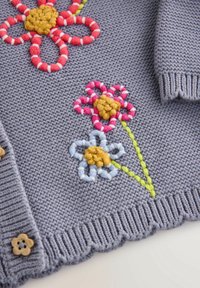 Tissu tricoté gris avec des fleurs brodées colorées en rouge, rose, bleu et jaune, avec des tiges vertes et des boutons en bois en forme de fleur.