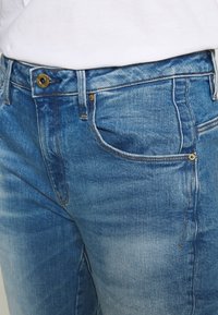 Gros plan sur la taille et la partie supérieure de la cuisse d'un jean en denim bleu avec un bouton en laiton et des rivets, porté avec une chemise blanche rentrée.