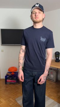 Marineblaue T-Shirt mit kurzen Ärmeln und gedrucktem Logo, kombiniert mit dunkelblauen, weit geschnittenen Jeans. Sichtbare Tätowierungen an den Armen.