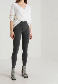 Weißer, gestrickter V-Ausschnitt Pullover, hochgeschnittene graue Skinny Jeans und Ankle Boots mit Schlangendruck und Blockabsatz. Glatte Texturen und taillierte Passform.