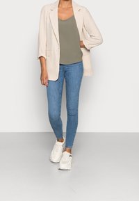 Donna che indossa jeans skinny blu, top verde oliva, blazer a quadri beige e sneakers bianche chunky, in piedi su uno sfondo semplice.