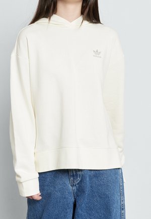 Osoba nosząca luźną, kremowo-białą bluzę Adidas z małym logo na piersi, w połączeniu z niebieskimi dżinsami na jasnym tle.