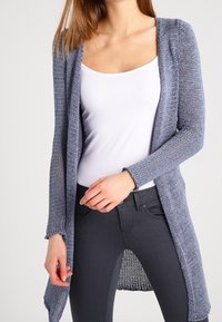 Lehké cardigan s dlouhým rukávem z texturovaného modrého úpletu. Otevřený přední design s delším zadním lemem a neupravenými švy.