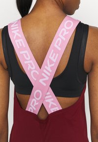 Mujer con sujetador deportivo negro y un mono burdeos con tirantes cruzados rosas etiquetados con "NIKE PRO" en la espalda.