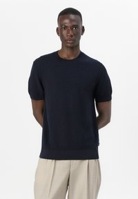 Mornarska pletena majica s kratkimi rokavi in krojem okrog vratu; teksturirano blago in rebrasti manšeti, v kombinaciji s svetlimi nabranimi hlačami.