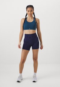 Sports-bh i teal, høyhåret lilla shorts med mørkt mønster, kombinert med hvite treningssko og synlige ankel-sokker.