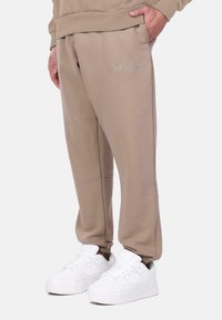 Pantalons de survêtement beige avec poches latérales et bords élastiques, arborant le texte brodé "Dropsize" sur le devant. Portés avec des baskets blanches.