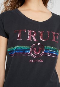 Woman wearing a black T-shirt with pink sequined "TRUE" text and multicolored sequined stripes below the text.