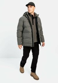 En pufferjakke i dæmpet grå med hætte, lagd over en mørk polo. Sorte bukser og lysebrune støvler fuldender outfittet.