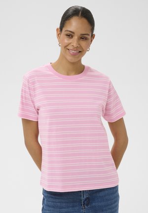 Femme souriante, portant un t-shirt à manches courtes rayé horizontalement rose et blanc, et un jean bleu, debout devant un fond clair uni.