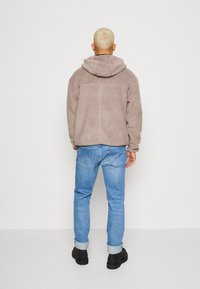 Giacca in fleece di un morbido taupe, con cappuccio, chiusura con zip e tessuto testurizzato. Indossata con jeans azzurri con risvolto e stivali neri.