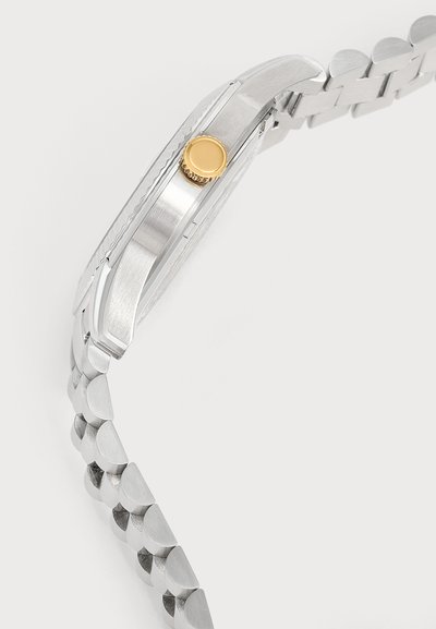 Gros plan d’un bracelet de montre en métal argenté avec une finition brossée et une couronne dorée sur le côté du boîtier de la montre.