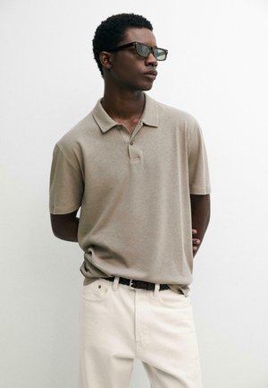 Uomo che indossa una polo beige a maniche corte, pantaloni bianchi, cintura nera intrecciata e occhiali da sole scuri, in piedi con le mani dietro la schiena.