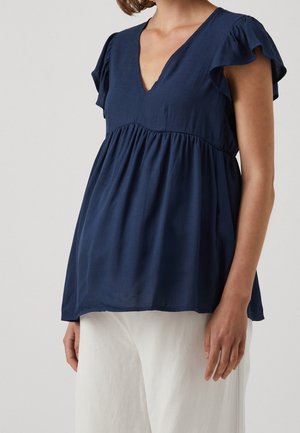 Femme portant un chemisier bleu marine à col en V avec des manches courtes volantées et un pantalon blanc ample sur un fond uni.