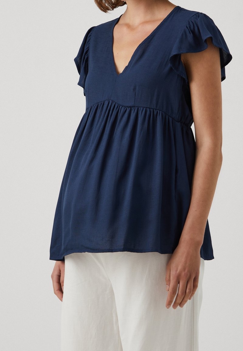 Femme portant un chemisier bleu marine à col en V avec des manches courtes volantées et un pantalon blanc ample sur un fond uni.